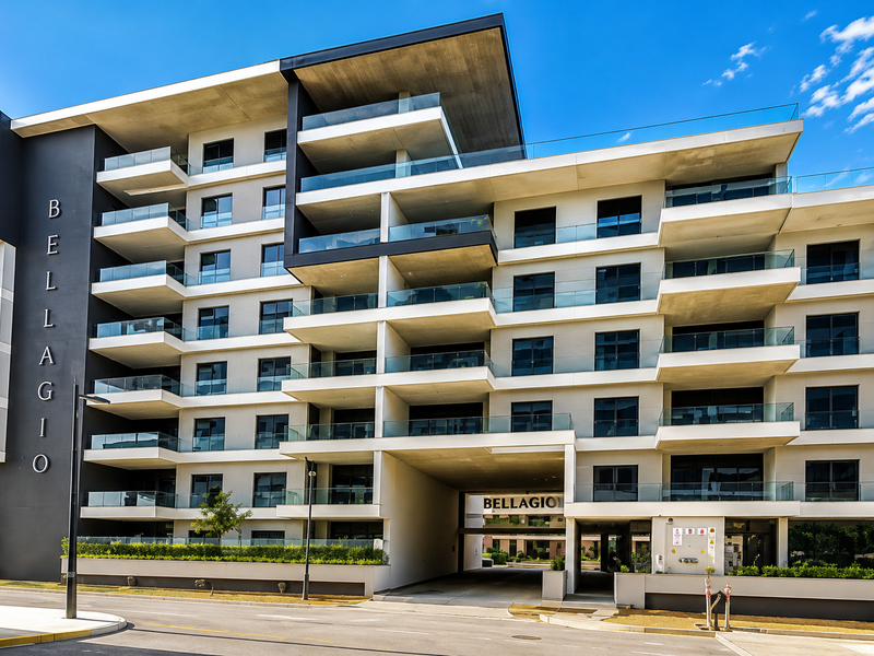 Modern multi-story apartment building facade featuring tiered balconies with glass balustrades and a covered ground-level driveway entrance.