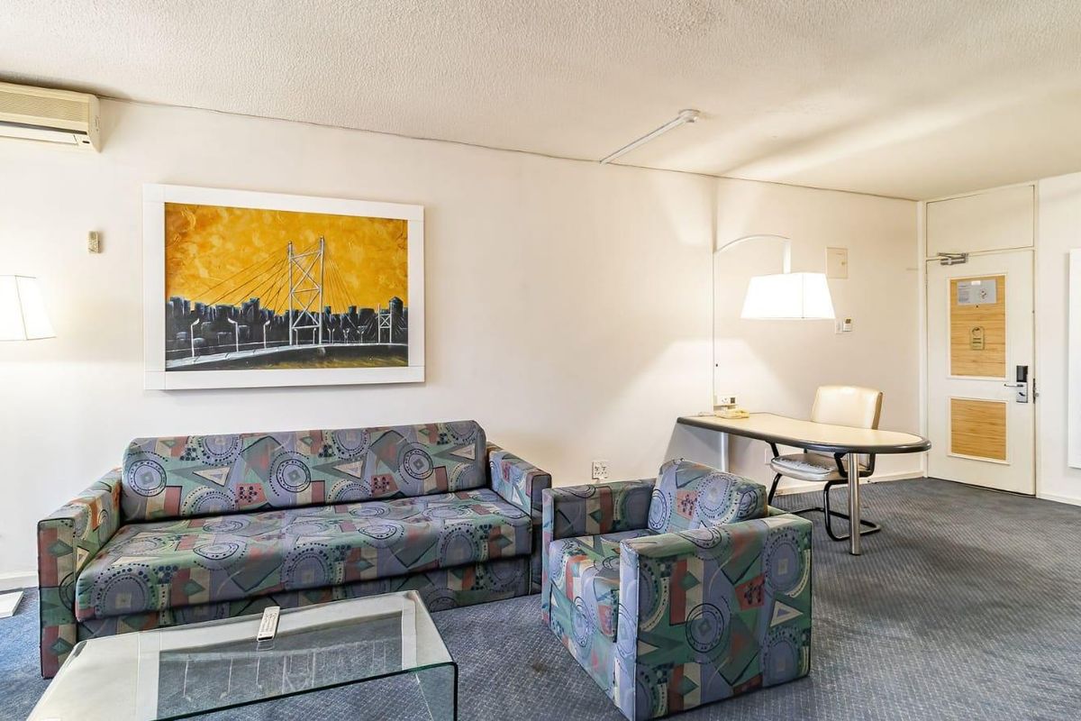 A dated living area featuring a built-in desk, wall-mounted air conditioning unit, and carpeted flooring, offering a functional space for relaxation or work.