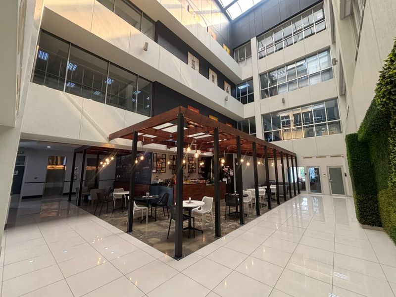 Modern office atrium featuring a central timber pergola seating area, high ceilings with skylights, polished tile flooring, and a decorative vertical garden wall.