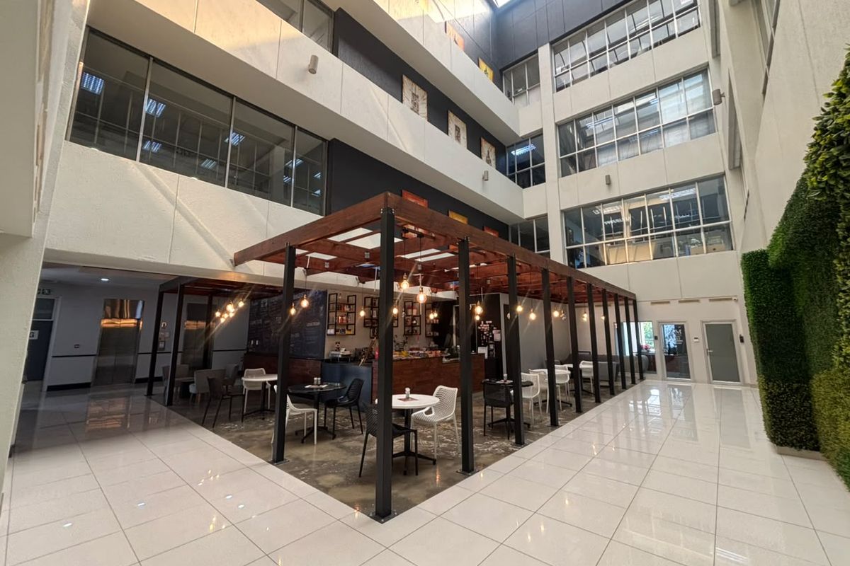Modern office atrium featuring a central timber pergola seating area, high ceilings with skylights, polished tile flooring, and a decorative vertical garden wall.