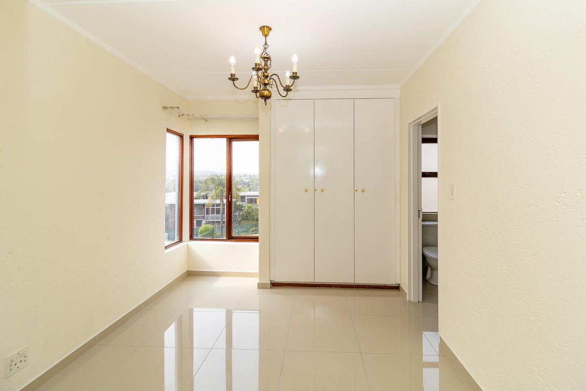 A bright, empty bedroom featuring tiled flooring, built-in cupboards, a classic chandelier, and a bay window with views.