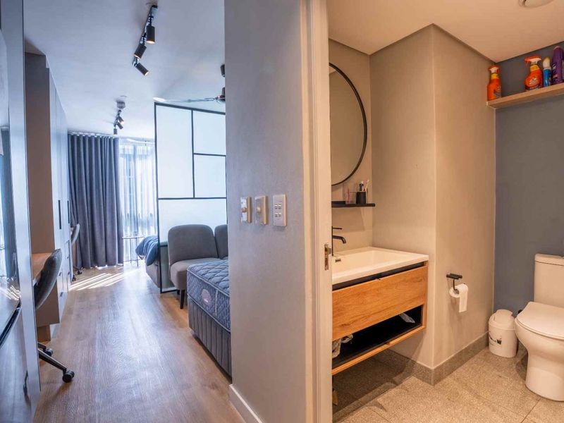 A modern bathroom featuring a wood-finish vanity, basin, and toilet, adjacent to an open-plan living and bedroom area with laminate flooring.