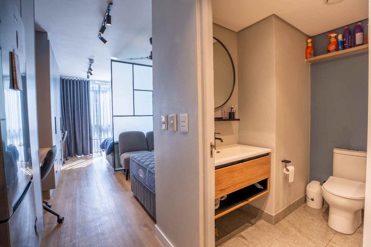 A modern bathroom featuring a wood-finish vanity, basin, and toilet, adjacent to an open-plan living and bedroom area with laminate flooring.