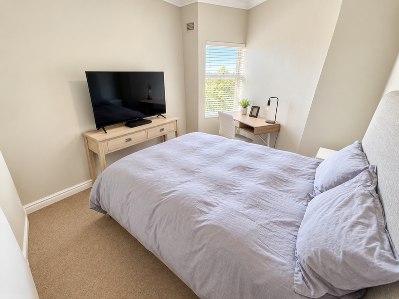 A well-lit bedroom featuring neutral wall tones, carpeted flooring, and a window with fitted venetian blinds.