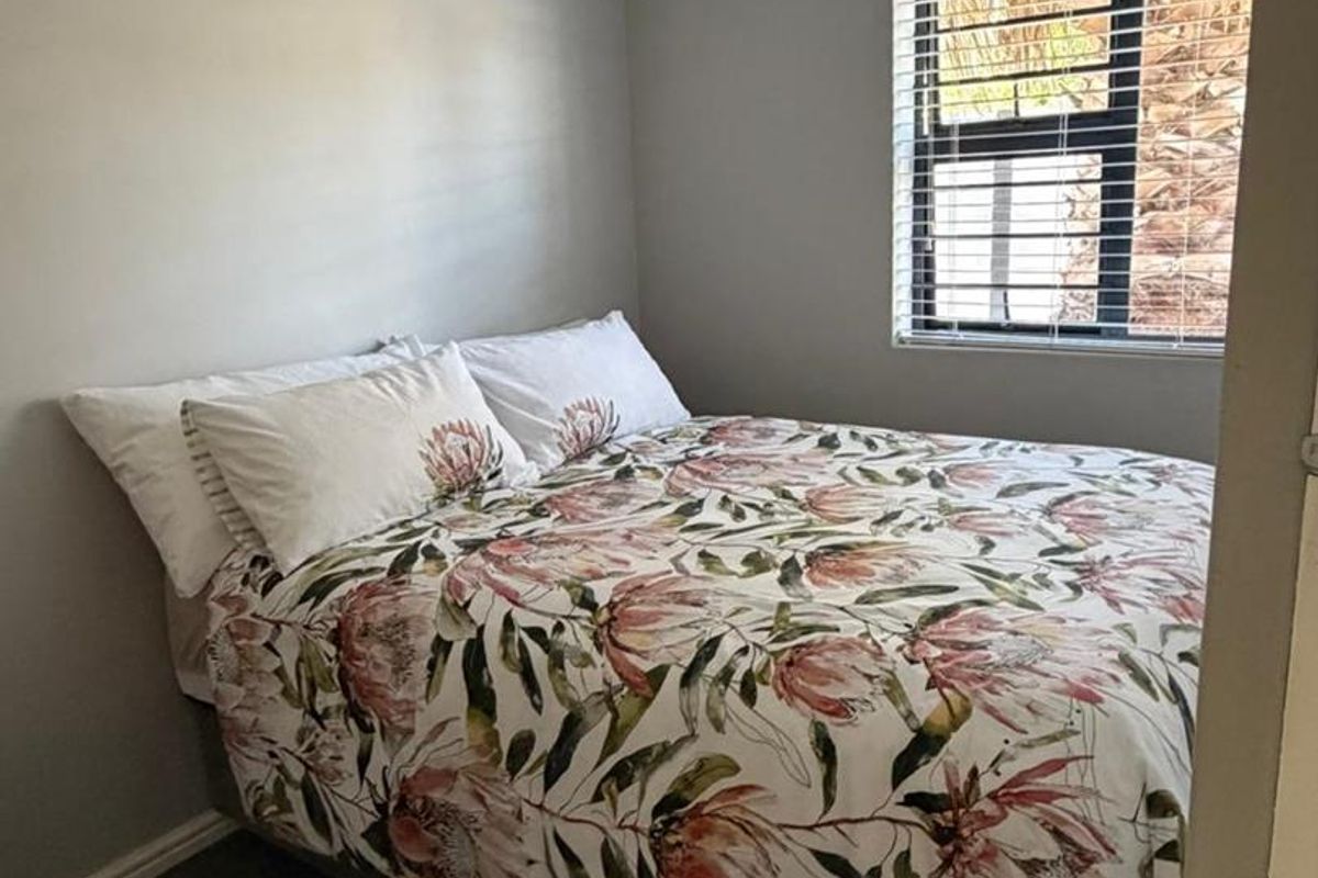 A compact bedroom featuring neutral grey walls, carpeted flooring, and a window fitted with venetian blinds.