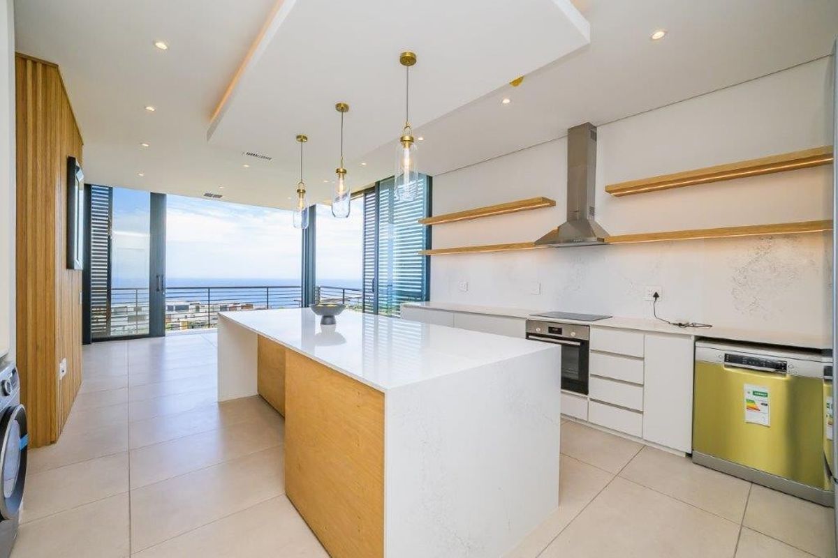 Modern open-plan kitchen featuring a central island, stone countertops, integrated appliances, floating shelves, and seamless access to a balcony with ocean views.