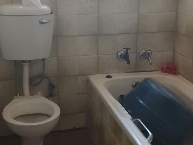 A dated bathroom featuring a standard toilet, bathtub with wall-mounted taps, and tiled walls with security bars on the window.