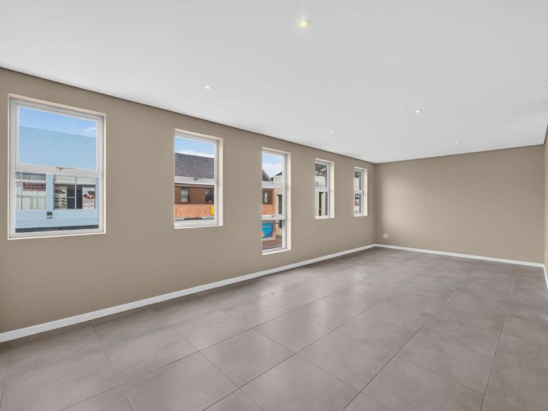 A bright, spacious, and empty living area featuring modern grey floor tiles, recessed downlights, and multiple windows providing natural light.