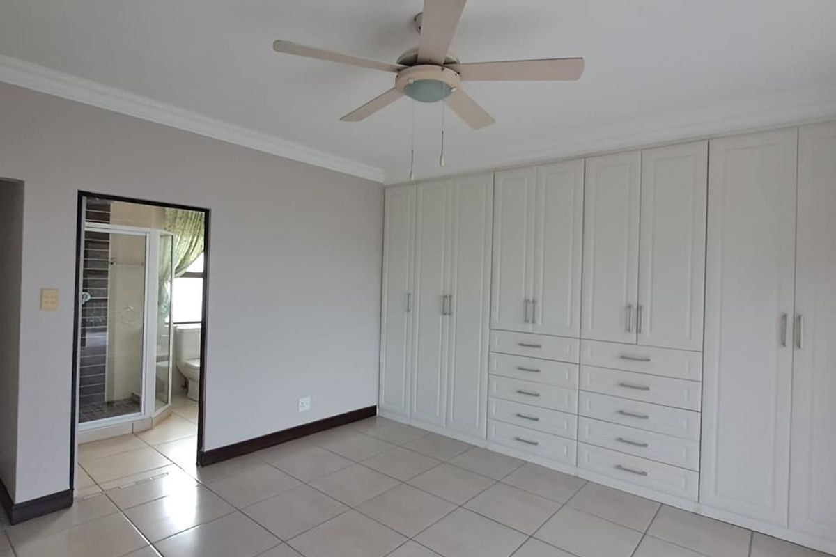 A spacious, empty bedroom featuring neutral tiled flooring, extensive built-in cabinetry, a ceiling fan, and direct access to an en-suite bathroom.