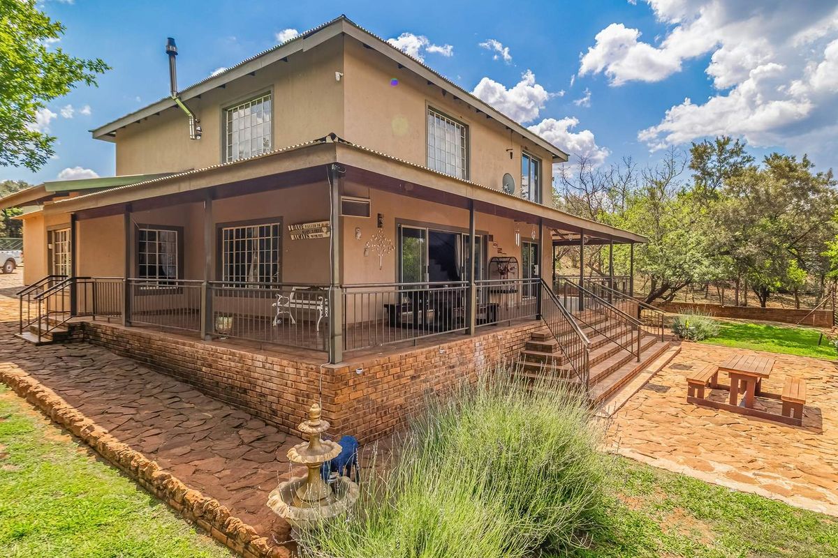 Rear view of a two-story farmhouse featuring a spacious covered patio, stone-paved terrace, and established garden surroundings.