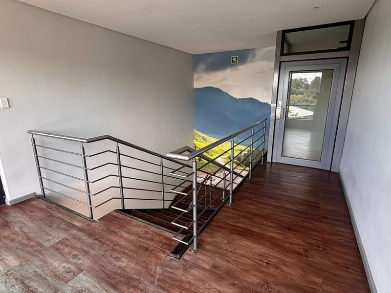 A modern office hallway featuring a staircase with a stainless steel balustrade, wood-look laminate flooring, and a glass-paneled door.