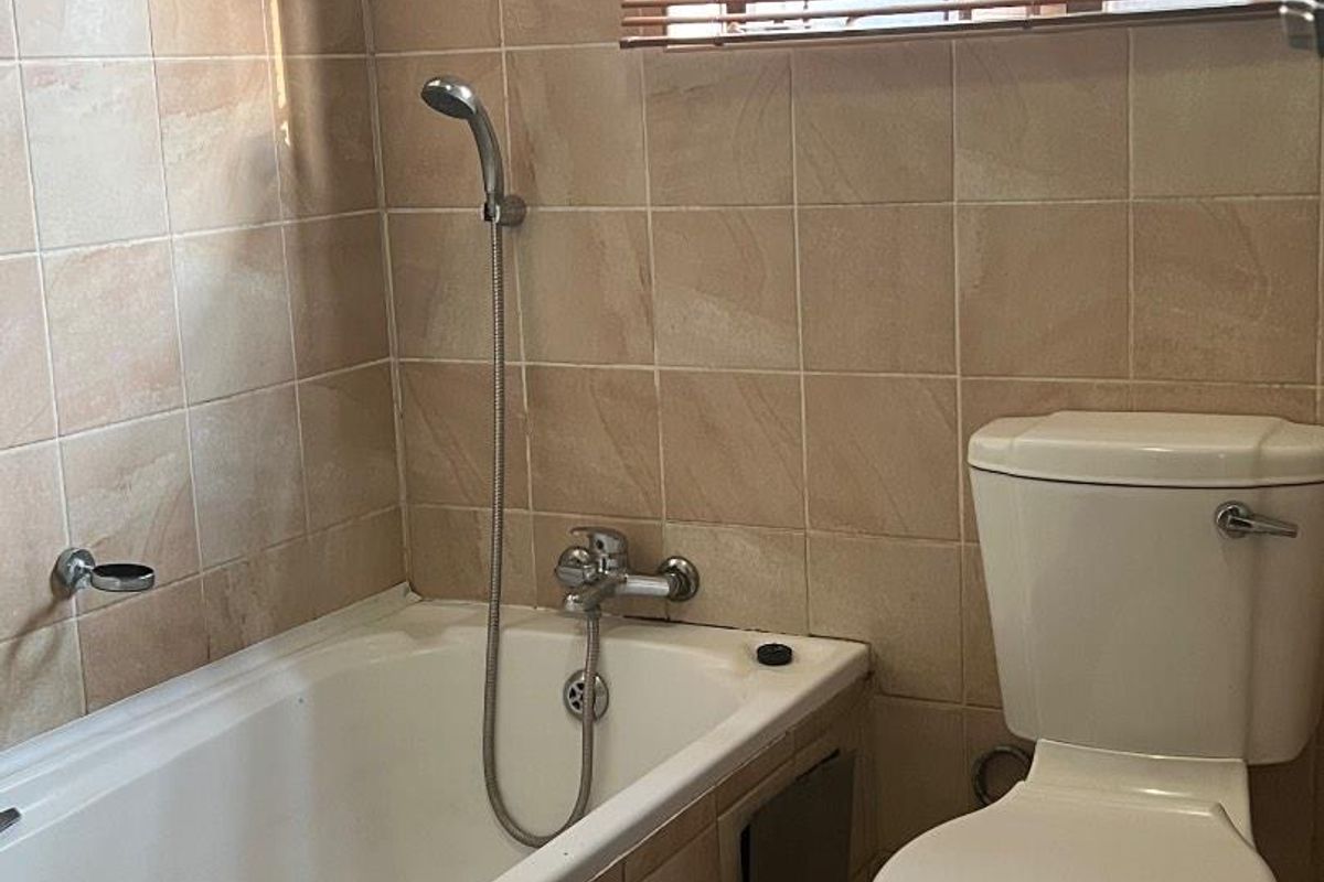 A functional bathroom featuring a built-in bathtub with a handheld shower mixer, a standard toilet, and neutral wall tiling.