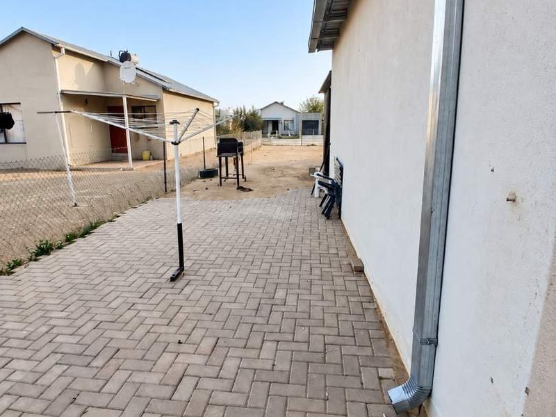 A rear exterior view showing a paved patio area alongside the house with a metal downpipe and perimeter fencing.