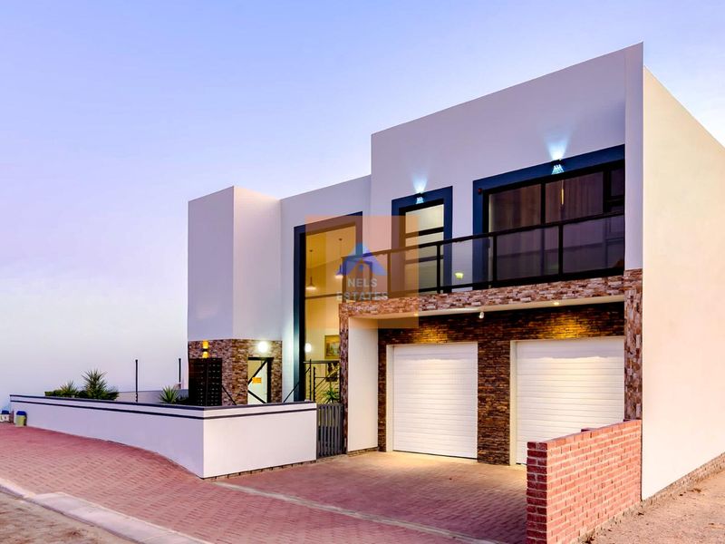 Modern double-story residential facade featuring a double garage, stone cladding accents, and an upper-level balcony with a glass balustrade.