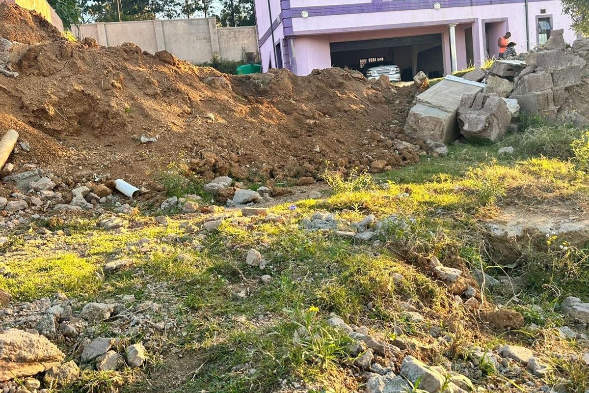 A view of a residential plot featuring uneven terrain, piles of excavated soil, and debris, with a neighboring property visible in the background.