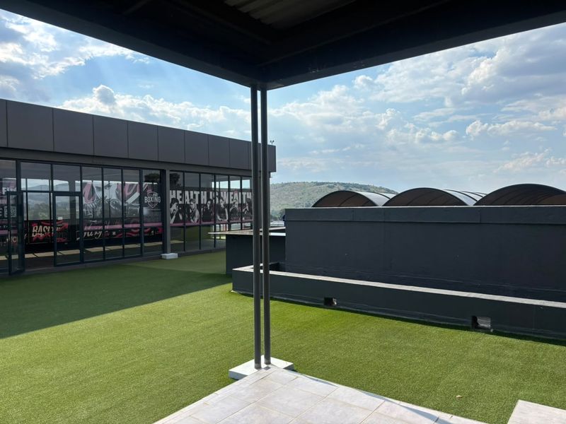 A spacious outdoor balcony area featuring artificial turf, metal structural supports, and a modern glass-fronted office space with scenic views.