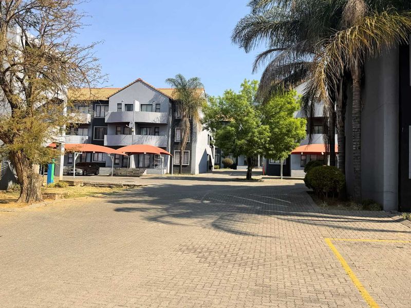Exterior view of a multi-story apartment complex featuring a paved driveway, established trees, and covered parking bays.