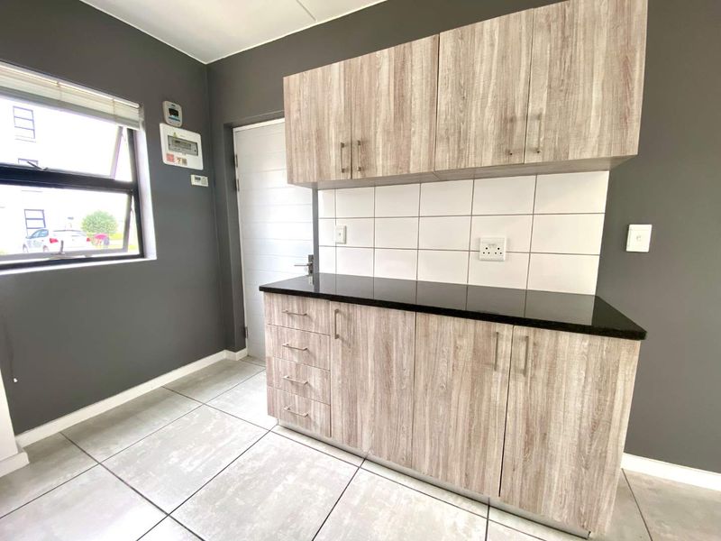 A clean, modern kitchen area featuring light wood-look cabinetry, a contrasting black granite countertop, white tiled backsplash, and neutral floor tiles.