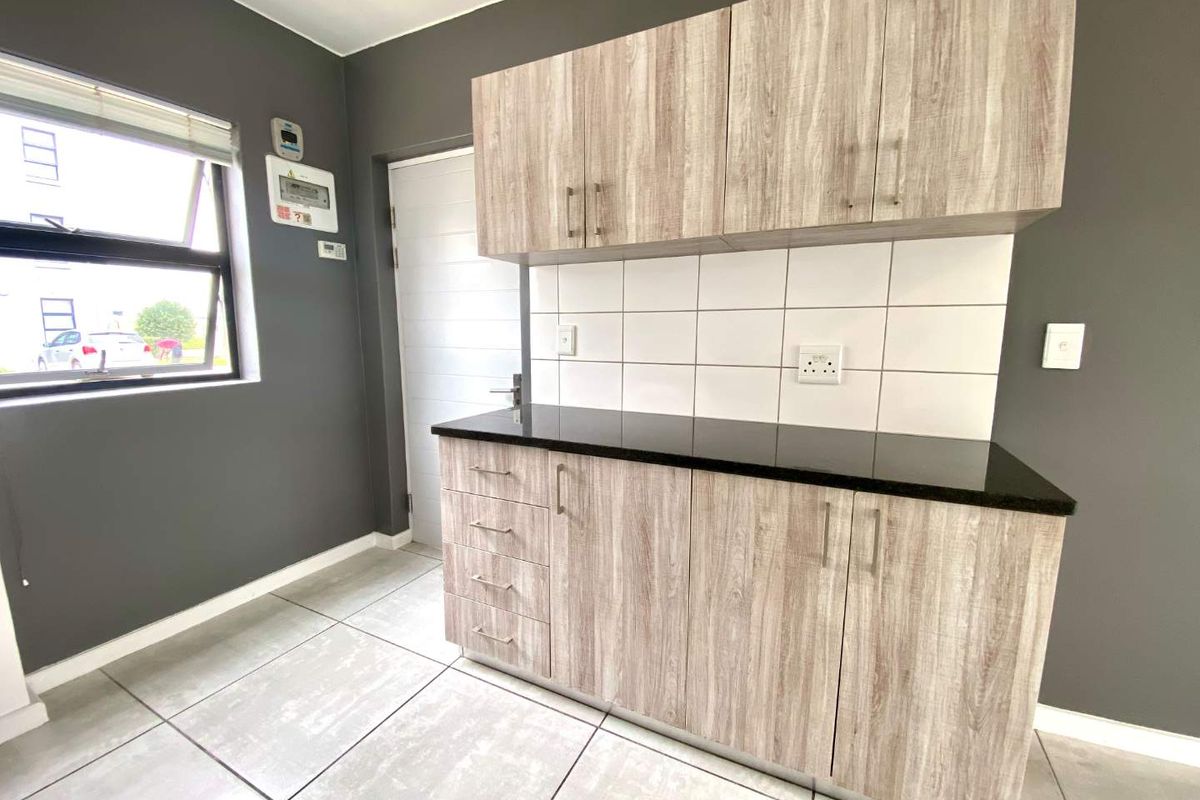A clean, modern kitchen area featuring light wood-look cabinetry, a contrasting black granite countertop, white tiled backsplash, and neutral floor tiles.