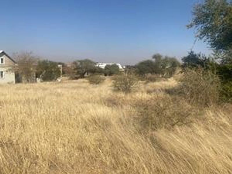 A view of a vacant residential plot featuring dry grass and native vegetation, with neighboring properties visible in the background.