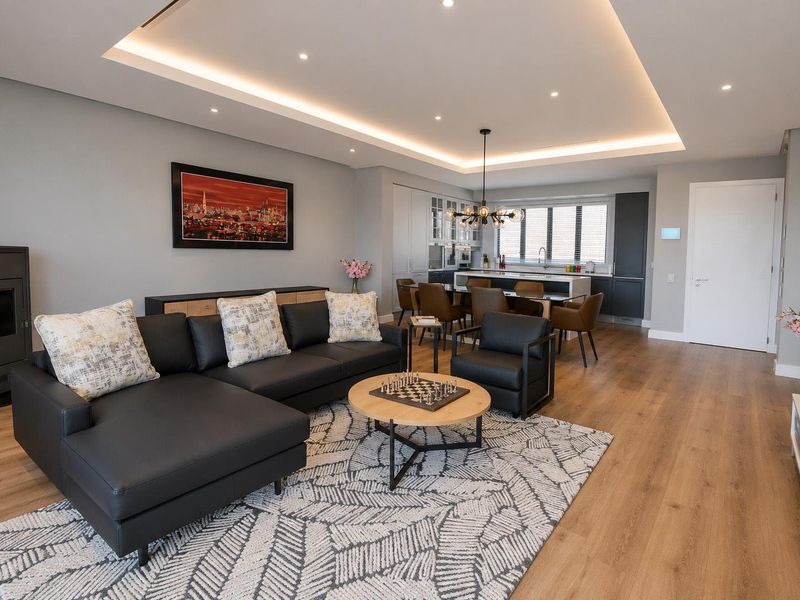 Modern open-plan living and kitchen area featuring recessed ceiling lighting, wood-look flooring, a sleek kitchen island, and a contemporary fireplace.