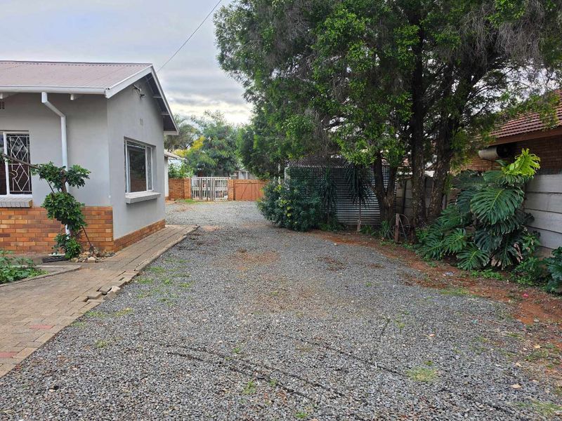 A view of the rear exterior area featuring a gravel driveway, brick paving, and perimeter security fencing with mature trees.