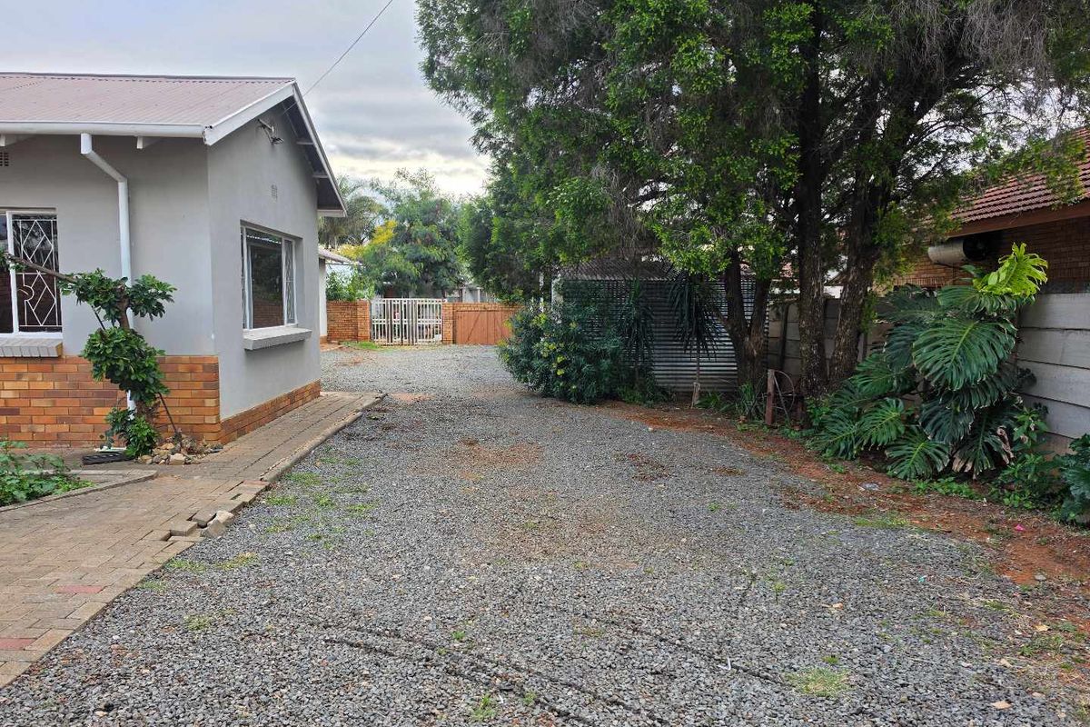 A view of the rear exterior area featuring a gravel driveway, brick paving, and perimeter security fencing with mature trees.