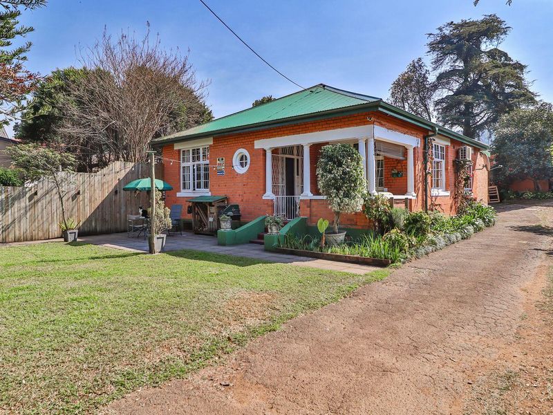 A charming single-story brick house featuring a covered front porch, a green metal roof, and a spacious front garden with a long driveway.