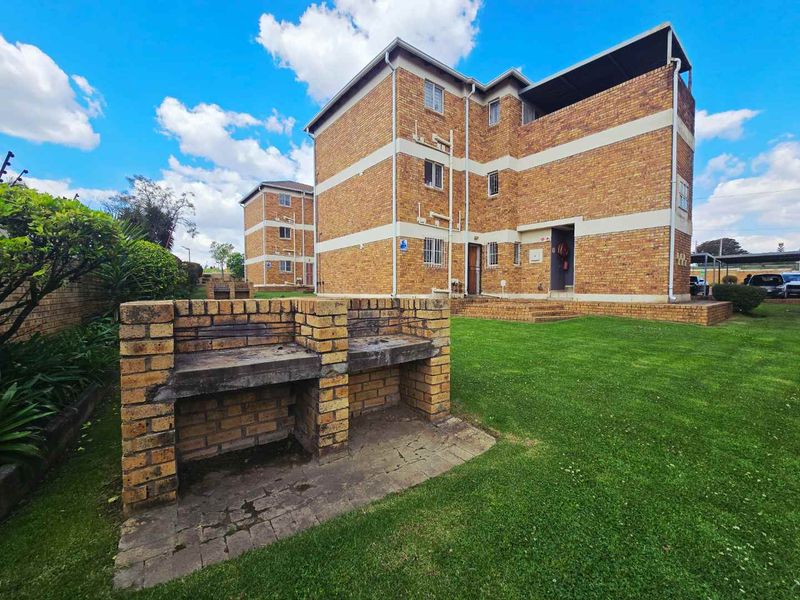 Exterior view of a brick apartment building featuring a communal lawn area and a built-in brick braai facility.