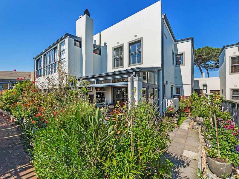 Rear view of the property featuring a paved walkway, lush garden beds, and an enclosed patio area with sliding glass doors.