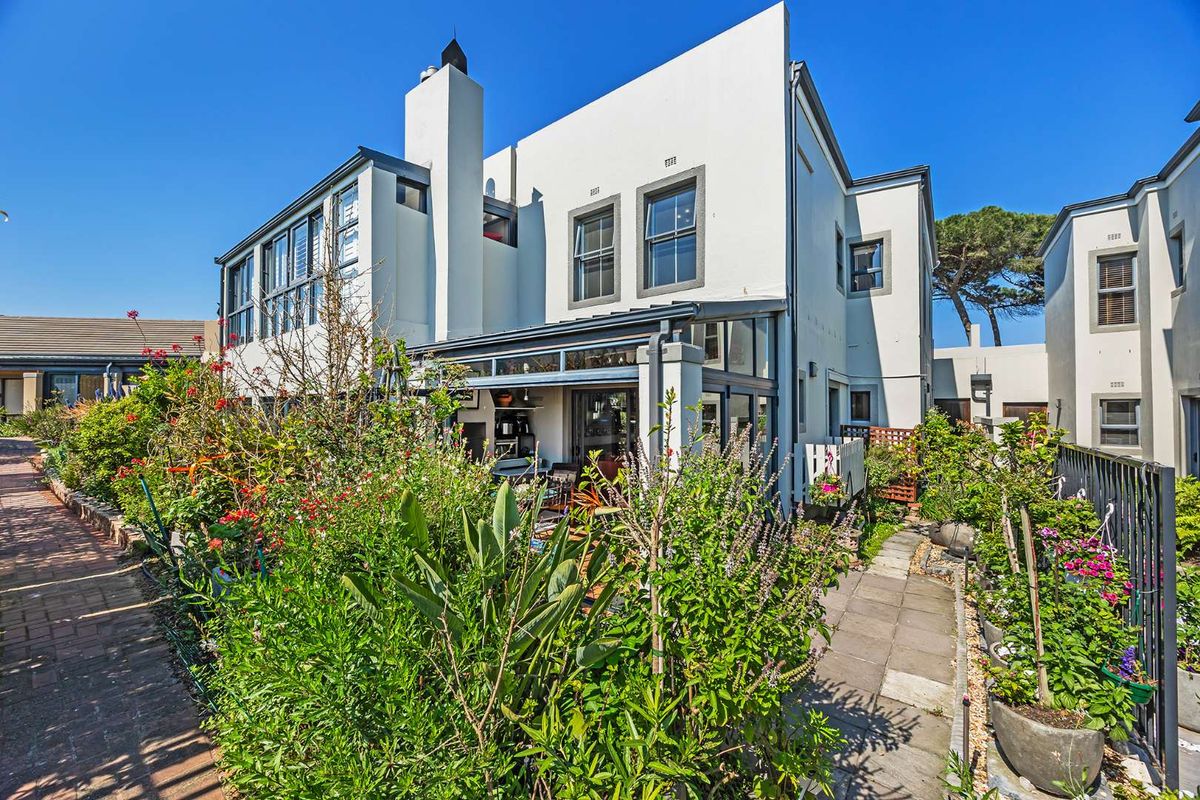 Rear view of the property featuring a paved walkway, lush garden beds, and an enclosed patio area with sliding glass doors.