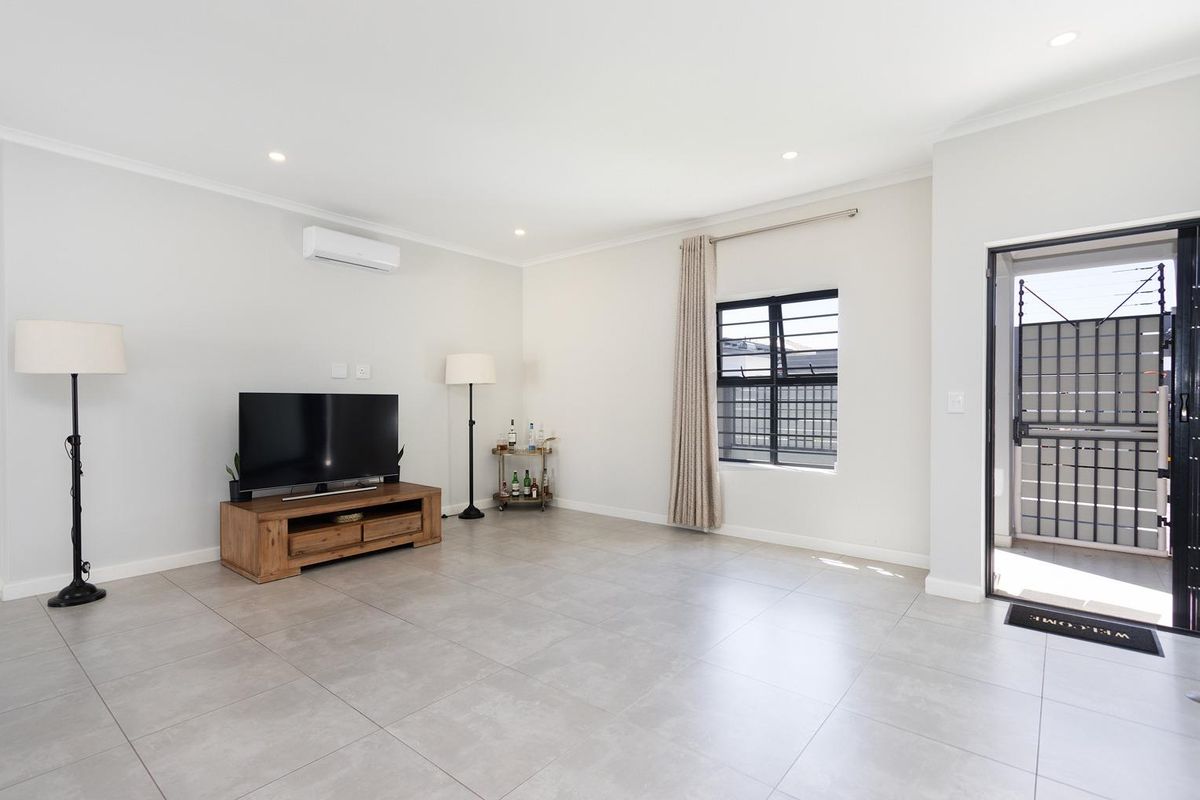A bright and spacious living room featuring neutral floor tiles, recessed lighting, a wall-mounted air conditioning unit, and direct access to the exterior.