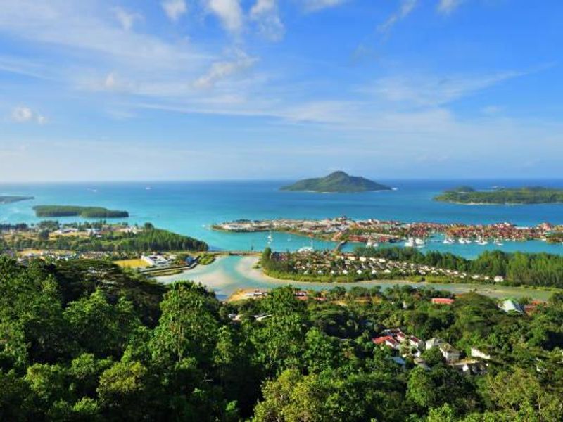 A scenic elevated view overlooking a coastal landscape, featuring lush greenery, a turquoise bay, and distant islands under a clear blue sky.
