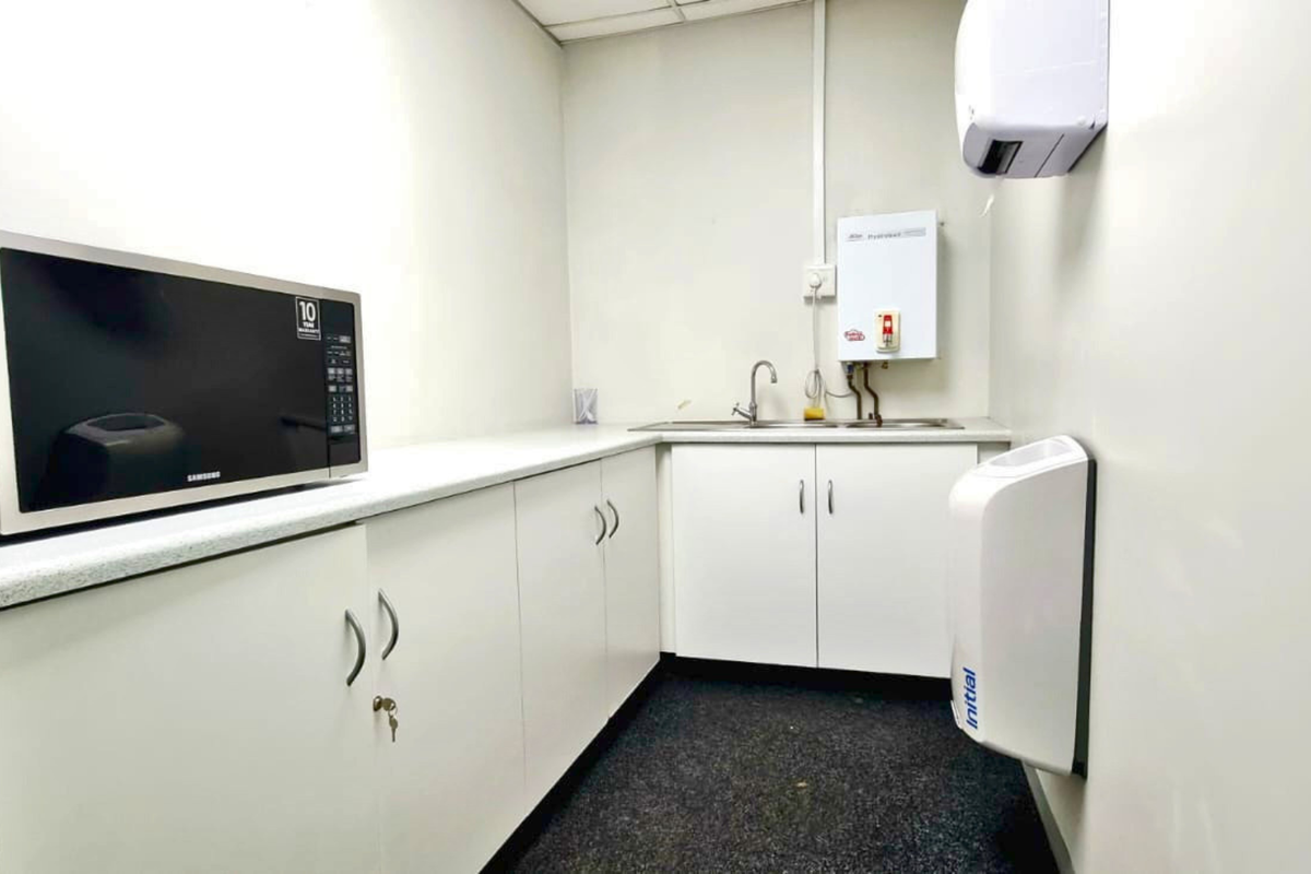 A functional office kitchenette featuring white built-in cabinetry, a stainless steel sink, an instant hot water heater, and wall-mounted utility dispensers.