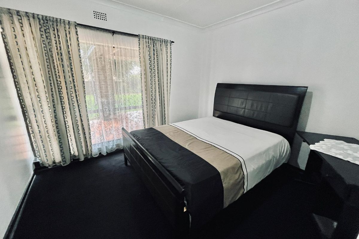 A well-lit bedroom featuring dark carpeting, neutral walls, and a large window with curtains providing natural light and access to the exterior.