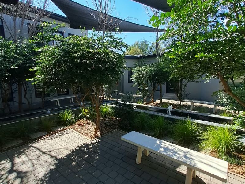 A well-maintained outdoor courtyard featuring paved areas, mature trees, a water feature, and overhead shade sails for a comfortable office break space.