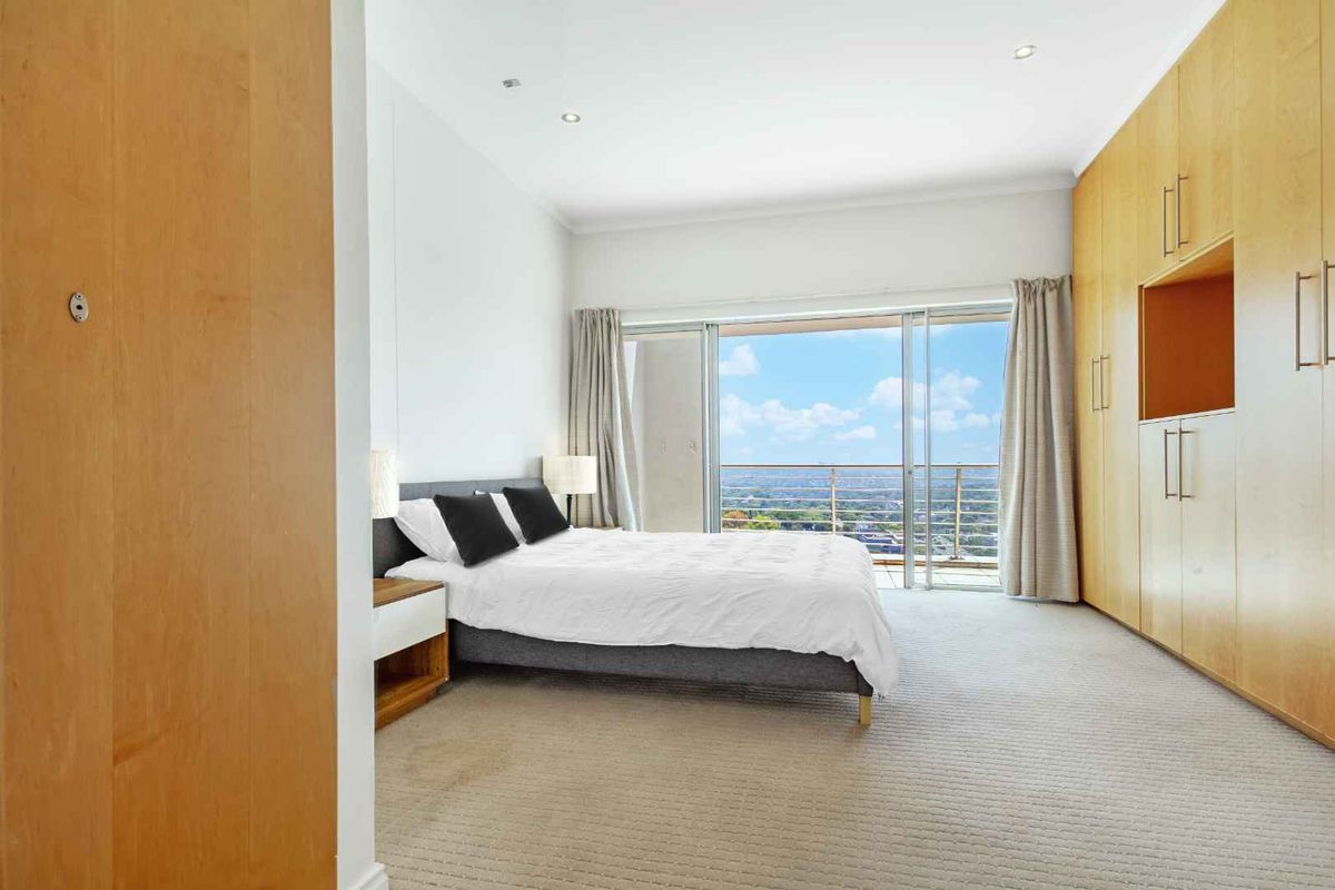 Spacious bedroom featuring extensive built-in wooden cabinetry, neutral carpeting, and large sliding glass doors opening onto a private balcony with expansive city views.