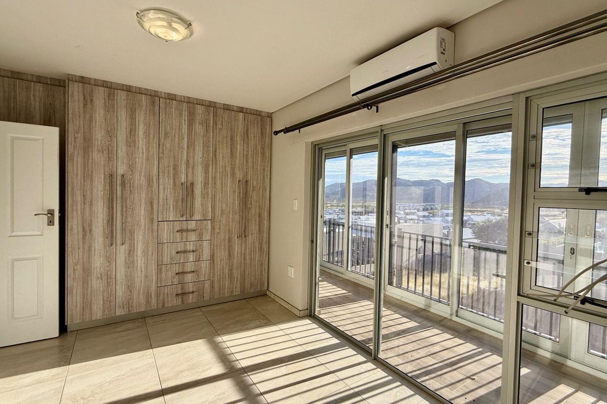 A bright bedroom featuring built-in wood-grain cabinetry, tiled flooring, a wall-mounted air conditioning unit, and large sliding glass doors opening to a balcony.