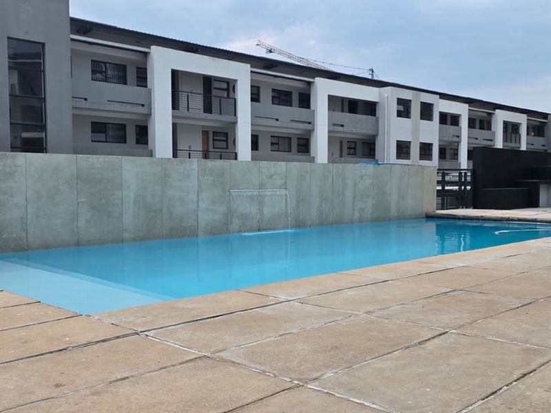 Communal swimming pool area featuring a modern concrete water feature wall, paved decking, and built-in braai facilities within an apartment complex.