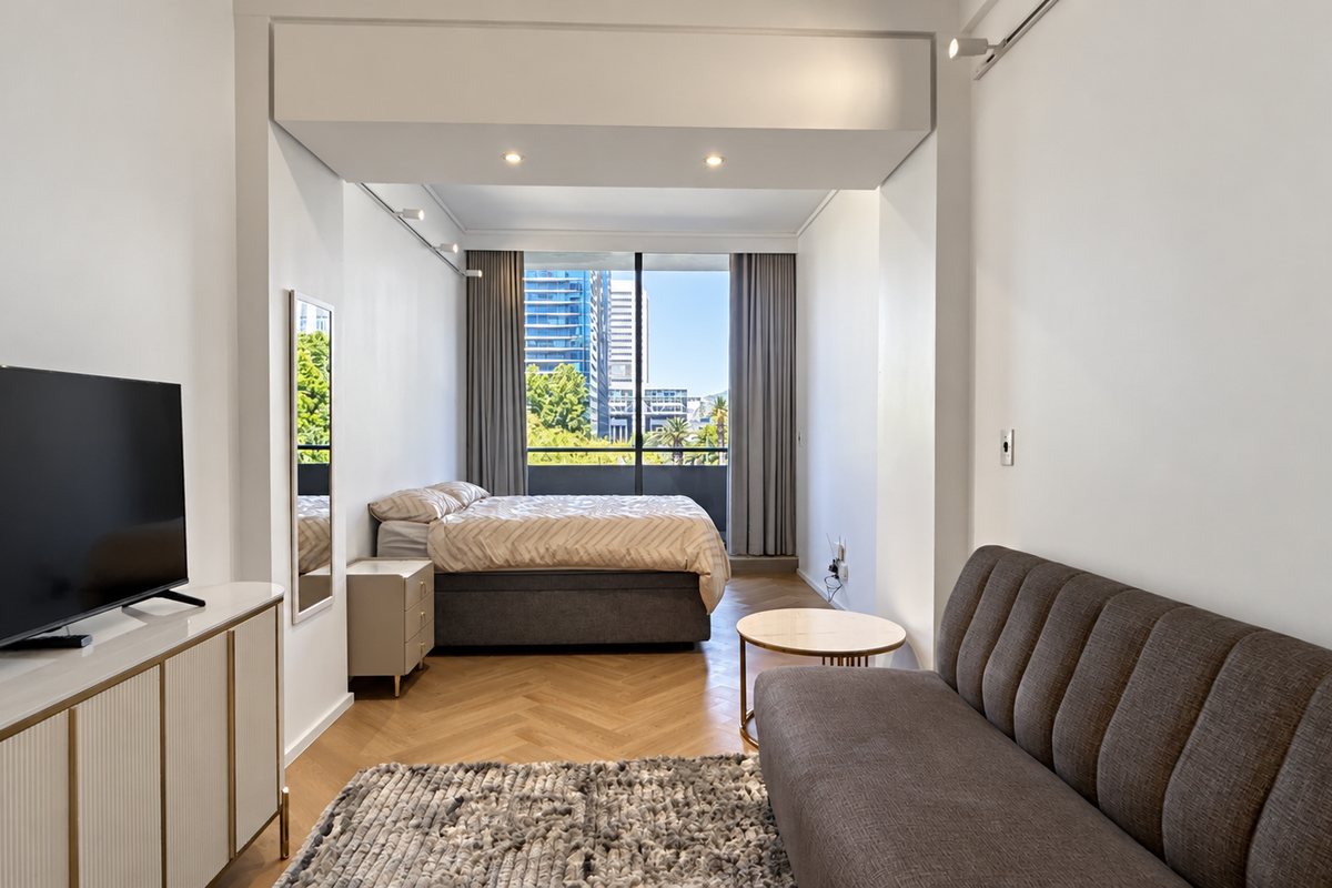 A modern open-plan studio apartment featuring elegant herringbone timber flooring, recessed lighting, and a large window providing natural light and urban views.