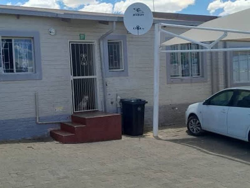 Front exterior view of the apartment featuring a paved driveway, carport, and security gate entrance.