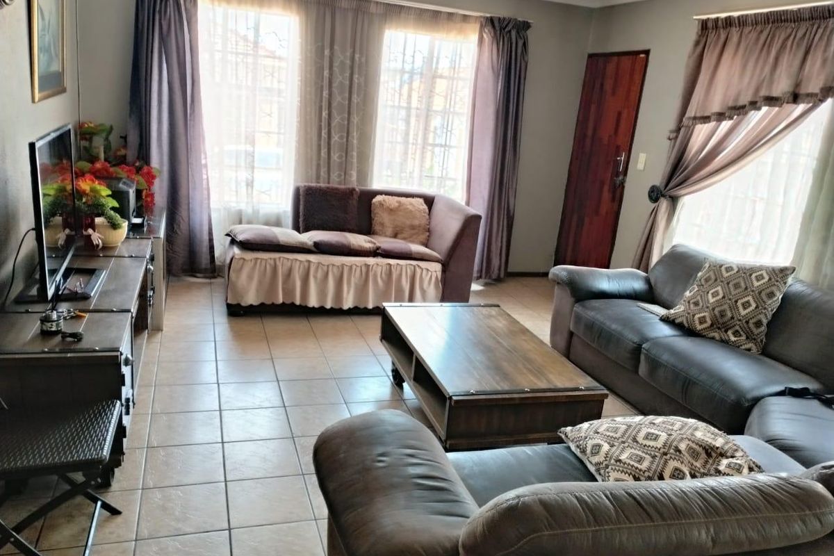 A spacious living room featuring neutral tiled flooring, large windows with curtains, and a solid wood door leading to the exterior.