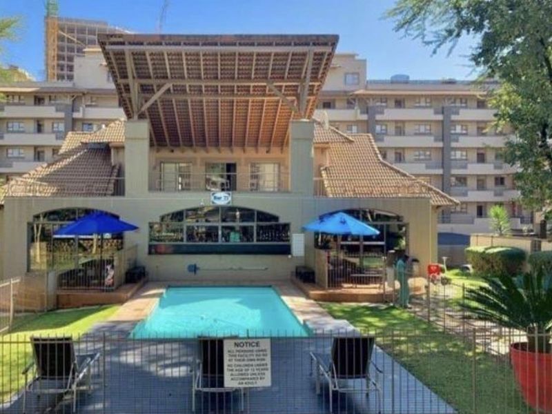 A communal pool area featuring a swimming pool, covered patio space, and landscaped garden surroundings within an apartment complex.
