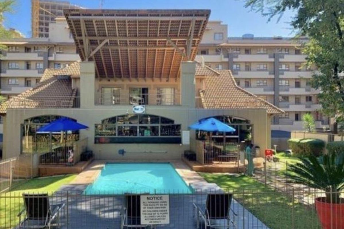 A communal pool area featuring a swimming pool, covered patio space, and landscaped garden surroundings within an apartment complex.