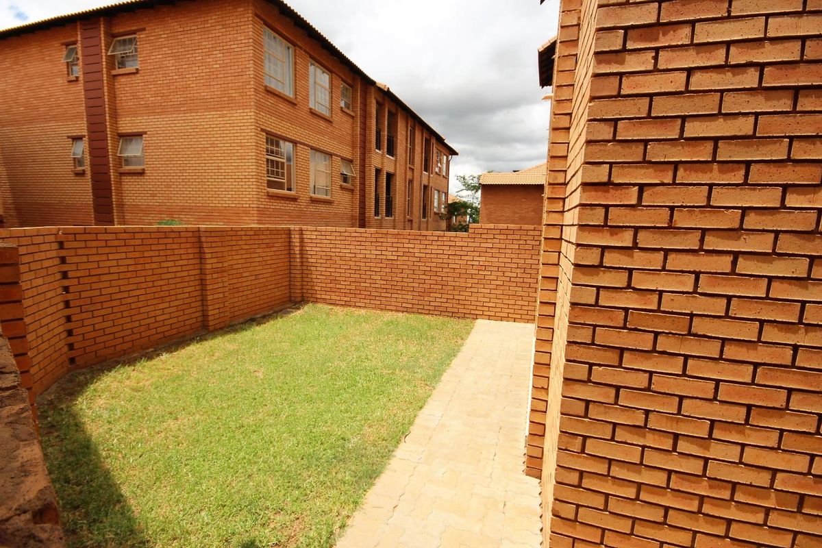 A private rear garden area featuring a paved walkway, established lawn, and enclosed by face brick boundary walls within an apartment complex.