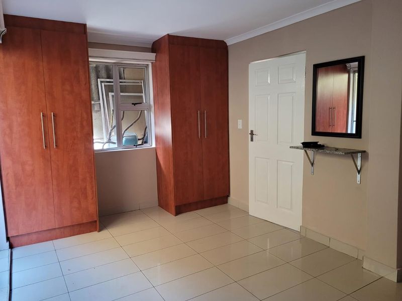 A clean, empty bedroom featuring built-in wooden cupboards, neutral tiled flooring, and a wall-mounted shelf, offering a functional and well-maintained living space.