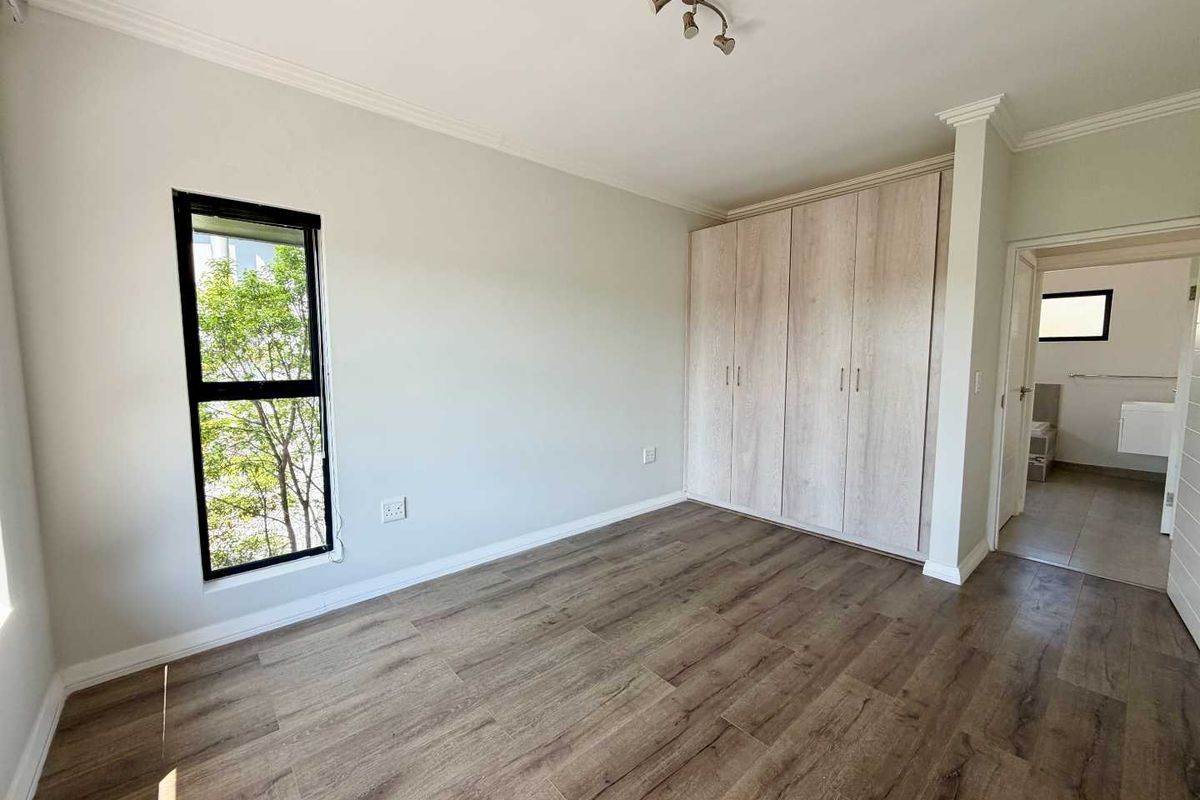 A bright, empty bedroom featuring modern laminate flooring, built-in cupboards, and a view through a vertical window, with an adjacent en-suite bathroom.