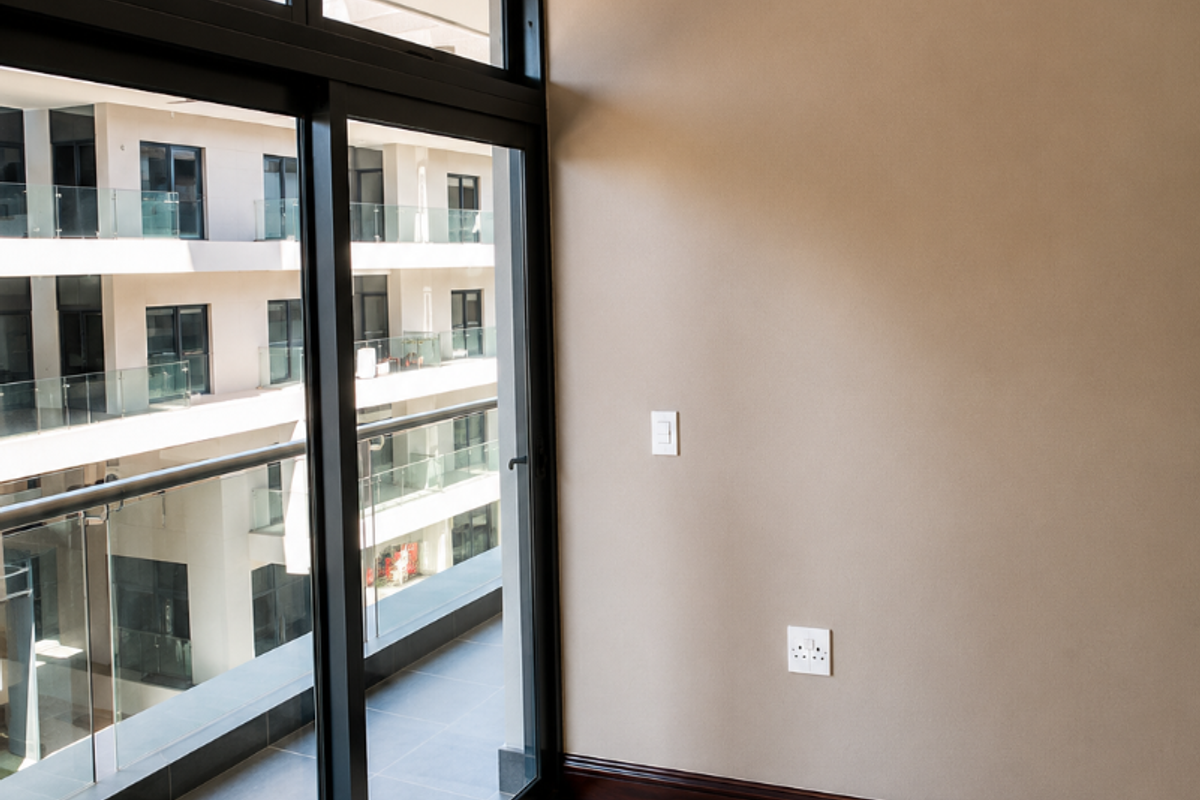 A bright, empty living area featuring large sliding glass doors that open onto a balcony, with neutral walls, tiled floors, and modern recessed ceiling details.