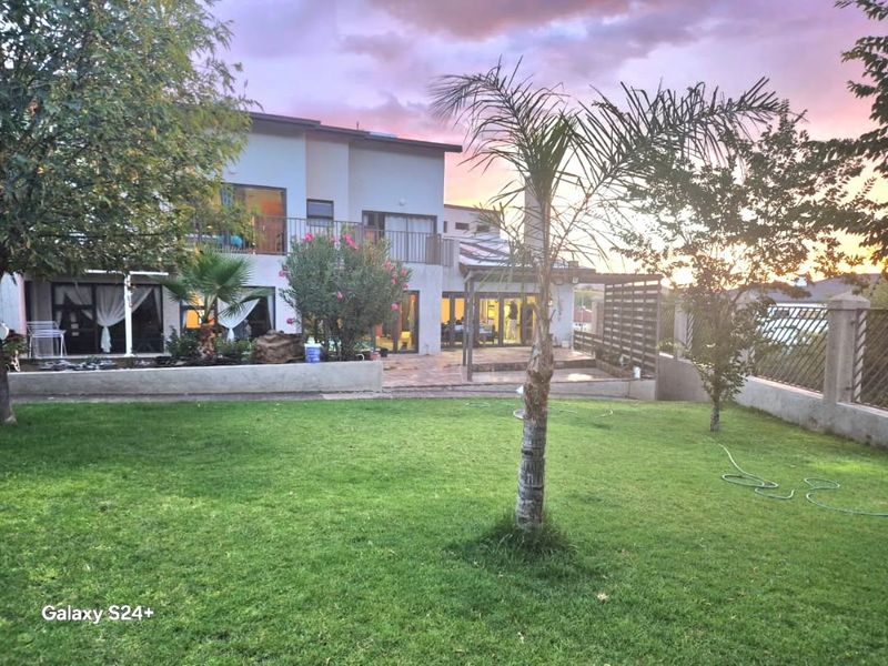Rear view of the property featuring a landscaped garden, paved patio area, pergola, and an upper-level balcony at sunset.