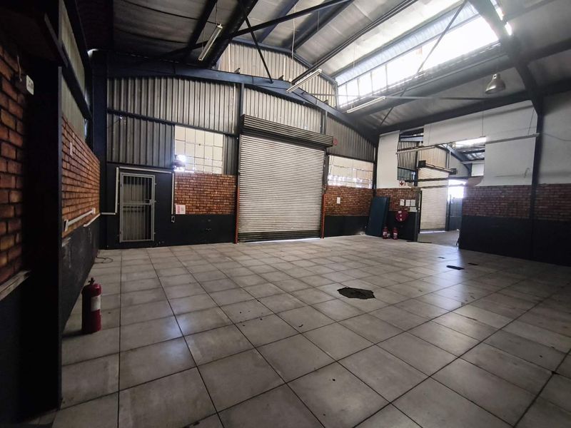 Spacious industrial utility area featuring high ceilings with exposed steel trusses, durable tiled flooring, facebrick accents, and a large roller shutter door for access.